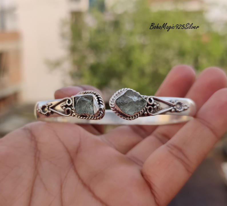 Puede incluir: Un brazalete de plata con dos piedras de aguamarina verde engastadas en el centro. El brazalete es abierto y ajustable.