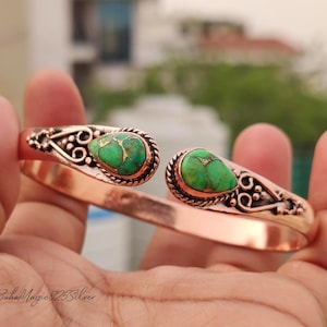 May include: Copper cuff bracelet with two teardrop-shaped, green turquoise stones. The bracelet features intricate, decorative metalwork around the stones and along the open ends. The bracelet is held in a hand, showcasing its design and color.