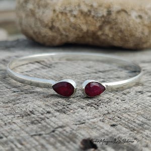 May include: A silver cuff bracelet with two teardrop-shaped, deep red gemstones. The open-ended bracelet sits on a weathered, grey wooden surface. The gemstones are set in silver bezels.
