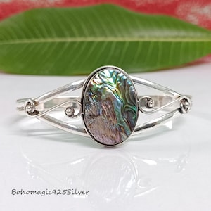 May include: A silver bracelet with a large oval abalone shell inlay. The bracelet has a delicate, intricate design with swirls and curves.