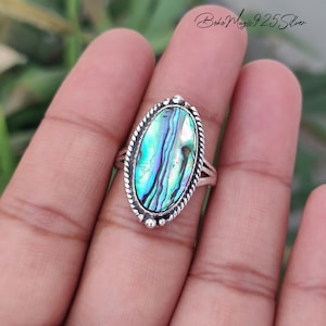 May include: An oval-shaped ring featuring an iridescent abalone shell inlay with vibrant green and blue tones. The stone is set in a polished silver frame with a twisted rope border and small bead accents. The text Boho Magic 925 Silver is visible.