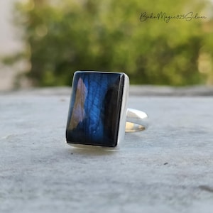 May include: A silver ring with a rectangular labradorite gemstone, displaying blue and black iridescence. The ring has a polished silver band and bezel setting. The text "BohoMagic925Silver" is visible in the background.