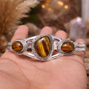 May include: Silver-toned cuff bracelet featuring three tiger's eye stones. The central stone is an irregular shape, flanked by two round stones. The stones display a warm, golden-brown color with striped patterns. The bracelet has a textured, braided design around the stones.