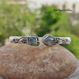 Puede incluir: Un brazalete de plata con dos acentos de piedras preciosas azul claro. El brazalete est&aacute; abierto y tiene un dise&ntilde;o delicado.