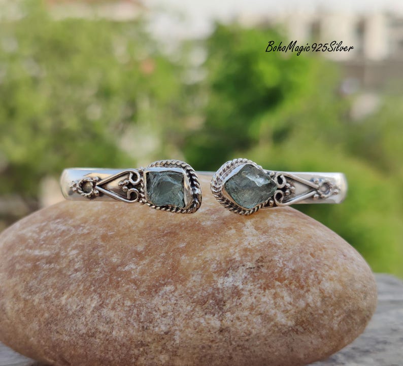 Puede incluir: Un brazalete de plata con dos acentos de piedras preciosas azul claro. El brazalete est&aacute; abierto y tiene un dise&ntilde;o delicado.