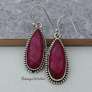Puede incluir: Un par de pendientes de plata con colgantes de piedras preciosas rojas en forma de lágrima. Los pendientes tienen un borde de cuentas alrededor de la piedra preciosa.
