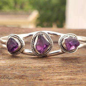 Peut inclure: Un bracelet manchette en argent orné de trois pierres d'améthyste brutes. Les pierres violettes sont serties dans des montures circulaires décoratives. Le bracelet est présenté sur une surface en bois.