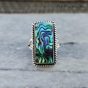 May include: A silver ring with a rectangular abalone shell inlay. The shell has a swirling pattern of blue, green, and purple.