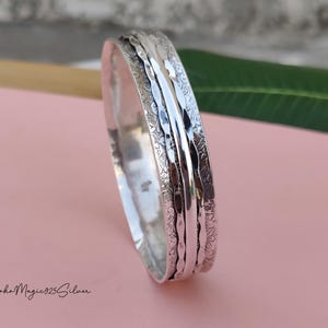 May include: A silver-colored bracelet with intricate floral and textured patterns. The bracelet has a wide band with multiple layers and a polished finish. The background is a soft pink with a green leaf and the text "BohoMagic925Silver".