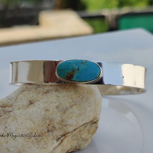 May include: A wide, polished silver cuff bracelet with an oval turquoise stone. The turquoise stone is set in a silver bezel. The bracelet is displayed on a light-coloured stone. The bracelet's width is approximately 2.5 cm.