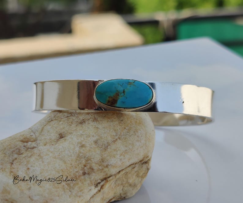 May include: A wide, polished silver cuff bracelet with an oval turquoise stone. The turquoise stone is set in a silver bezel. The bracelet is displayed on a light-coloured stone. The bracelet's width is approximately 2.5 cm.