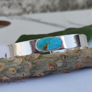 May include: A silver cuff bracelet featuring an oval turquoise stone. The turquoise stone has brown and blue hues. The bracelet rests on a textured wooden branch. The words "Boho Magic 925 Silver" are at the bottom.