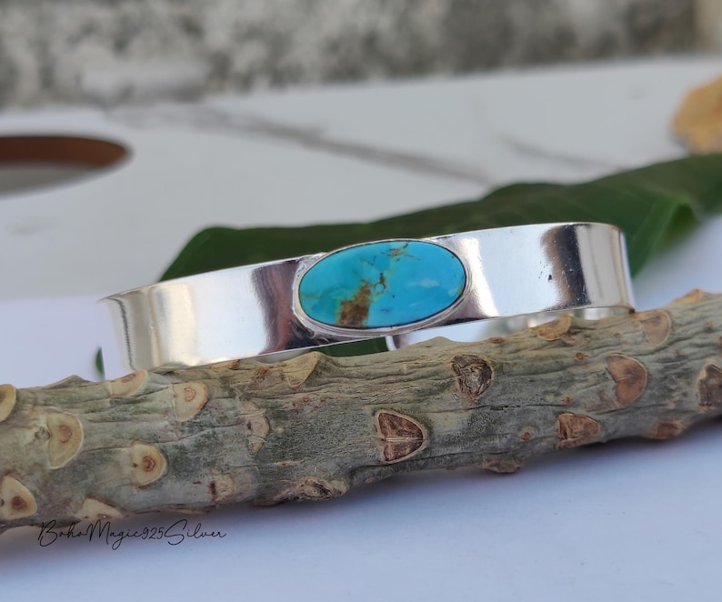 May include: A silver cuff bracelet featuring an oval turquoise stone. The turquoise stone has brown and blue hues. The bracelet rests on a textured wooden branch. The words "Boho Magic 925 Silver" are at the bottom.