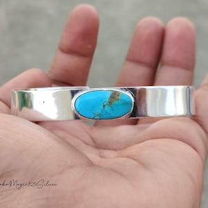 May include: A silver cuff bracelet with a bright turquoise stone. The oval turquoise stone is set in a polished silver band. The bracelet is displayed on a hand.