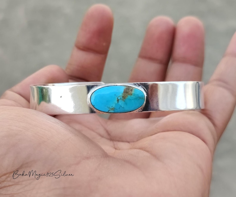 May include: A silver cuff bracelet with a bright turquoise stone. The oval turquoise stone is set in a polished silver band. The bracelet is displayed on a hand.