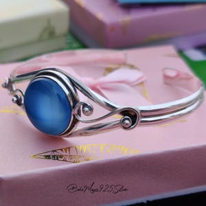 May include: A silver bangle bracelet with a blue cabochon gemstone. The bracelet has a delicate, ornate design with swirls and beads.