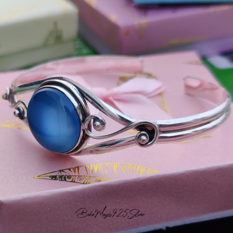 May include: A silver bangle bracelet with a blue cabochon gemstone. The bracelet has a delicate, ornate design with swirls and beads.