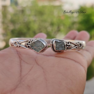 Puede incluir: Un brazalete de plata con dos acentos de piedras preciosas verdes. El brazalete tiene un dise&ntilde;o delicado y adornado y est&aacute; hecho de plata de ley 925.
