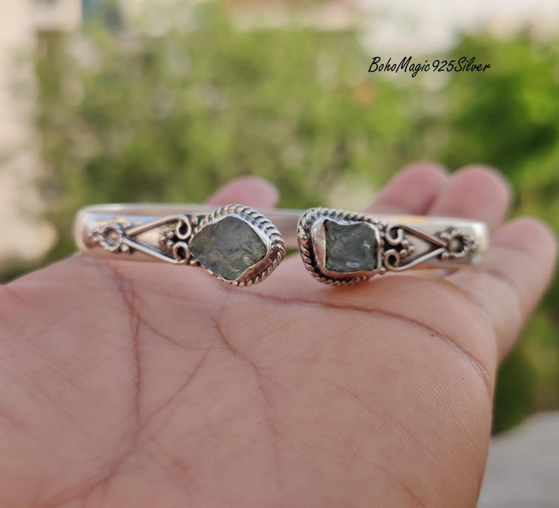 Puede incluir: Un brazalete de plata con dos acentos de piedras preciosas verdes. El brazalete tiene un dise&ntilde;o delicado y adornado y est&aacute; hecho de plata de ley 925.