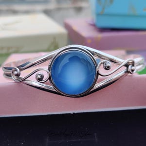 May include: A silver bangle bracelet with a large, round, light blue gemstone set in the center. The bracelet has a delicate, ornate design.