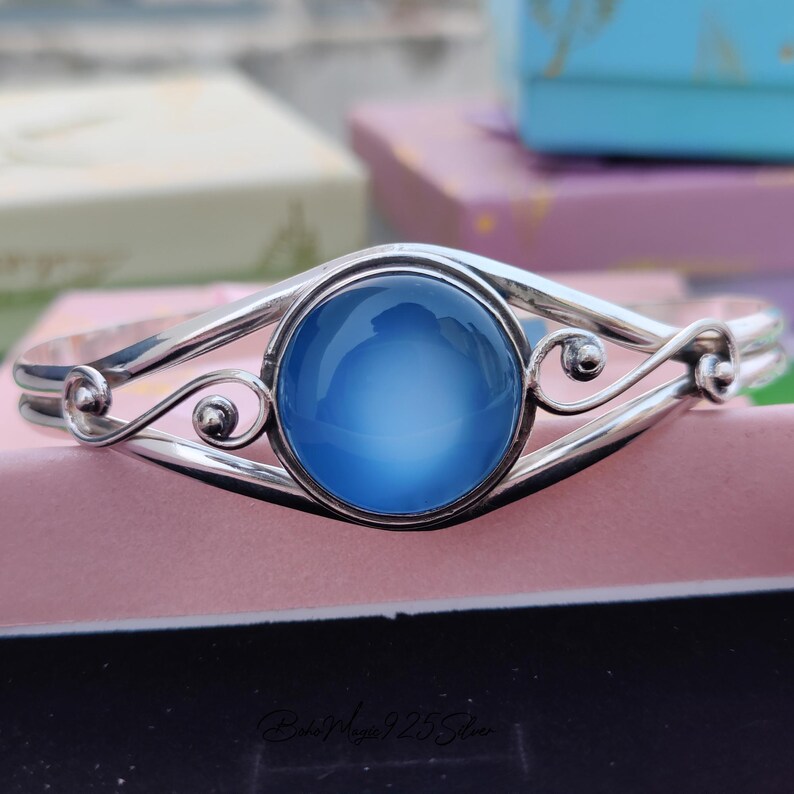 May include: A silver bangle bracelet with a large, round, light blue gemstone set in the center. The bracelet has a delicate, ornate design.