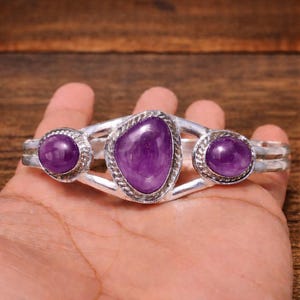 May include: Silver-toned cuff bracelet featuring three polished, oval-shaped purple gemstones. The central stone is larger and irregularly shaped, while the two flanking stones are smaller and round. The bracelet has a simple, elegant design.