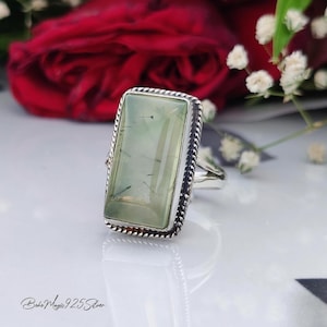 May include: A silver ring with a rectangular, light green gemstone. The gemstone has dark inclusions and is set in a detailed silver frame. The ring is on a white surface with a blurred background of red roses and white flowers. The text "Boho Magic 925 Silver" is at the bottom.