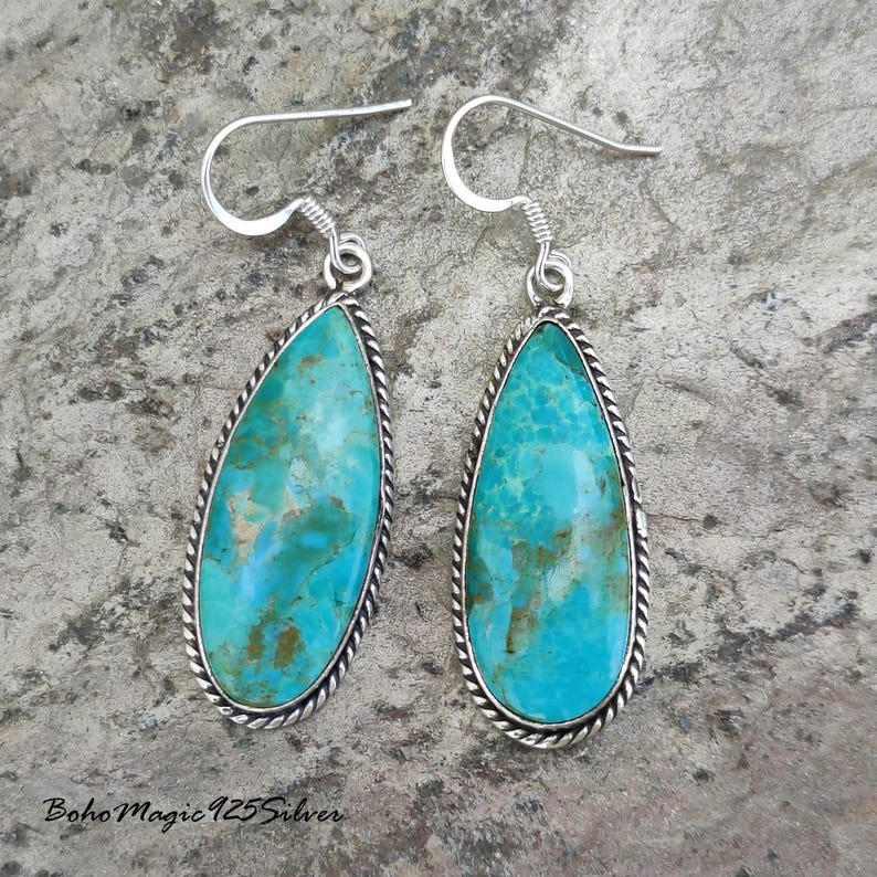 May include: A pair of turquoise teardrop earrings with a silver-colored rope-like border. The turquoise stones have a mottled appearance with shades of blue and brown. The earrings have a hook closure.