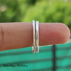 Puede incluir: Dos anillos de plata, uno liso y otro texturizado, se muestran en un dedo sobre un fondo verde y marrón borroso. Los anillos están juntos, mostrando su diseño y brillo. Los anillos probablemente son de plata de ley.