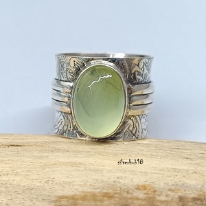 May include: A silver ring featuring a green gemstone set in a wide band. The band is decorated with a swirling pattern.