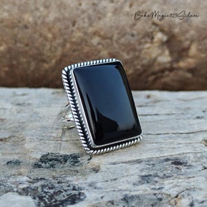 May include: A rectangular black onyx ring with a silver-colored, twisted rope design border. The ring has a large, polished black stone and a wide band. The ring is set on a natural, textured surface.