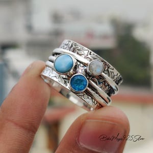 Puede incluir: Un anillo giratorio de plata con tres piedras preciosas azules y blancas. El anillo tiene un diseño floral y está puesto en un dedo.