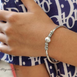 Peut inclure: Un bracelet en argent avec une perle décorative et un motif texturé. Le bracelet est porté au poignet.