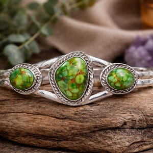 May include: A silver cuff bracelet with three green and brown stone settings. The central stone is oval-shaped, flanked by two round stones. The bracelet is displayed on a piece of wood.