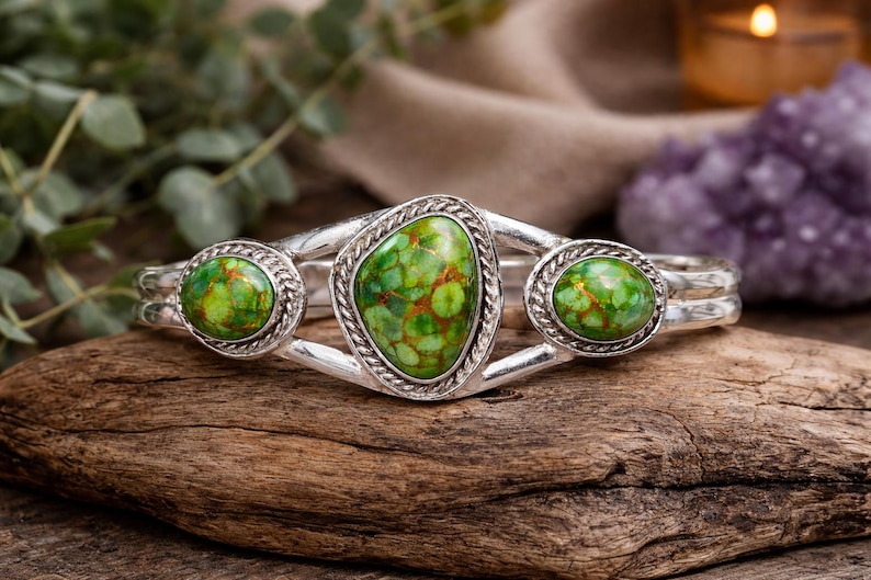 May include: A silver cuff bracelet with three green and brown stone settings. The central stone is oval-shaped, flanked by two round stones. The bracelet is displayed on a piece of wood.