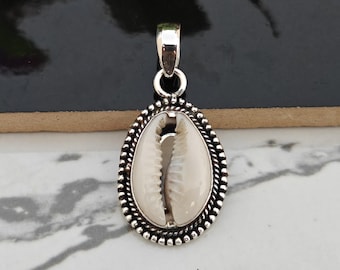 May include: A silver pendant featuring a white cowrie shell with a detailed silver border. The pendant is oval shaped and has a loop at the top for attaching to a chain.