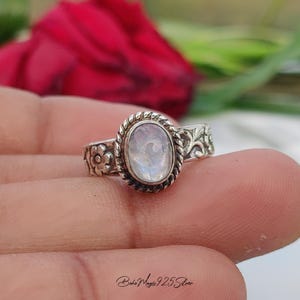 May include: A silver ring featuring an oval moonstone with an iridescent sheen. The ring has a braided border and floral and leaf patterns on the band. The ring is on a finger, with a red rose in the background.