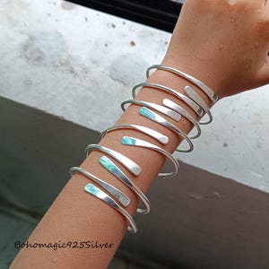 May include: A set of silver bangles with a hammered finish. The bangles are stacked on a person's arm and have a subtle turquoise hue.