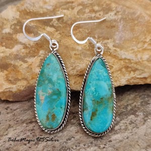 May include: A pair of turquoise teardrop earrings with a silver rope-like border and silver ear hooks. The turquoise stones have a mottled appearance with shades of blue and brown. The earrings are displayed on a textured, neutral-toned surface.