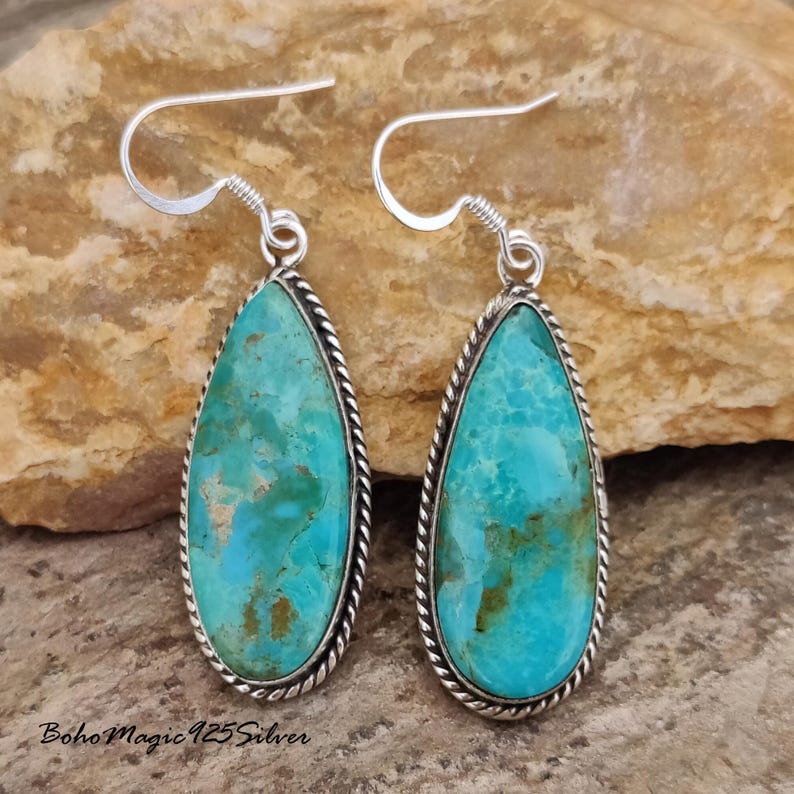 May include: A pair of turquoise teardrop earrings with a silver rope-like border and silver ear hooks. The turquoise stones have a mottled appearance with shades of blue and brown. The earrings are displayed on a textured, neutral-toned surface.
