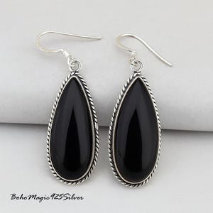 May include: A pair of teardrop-shaped earrings with polished black stones. The stones are framed by a silver-colored, twisted rope design. The earrings have a simple hook closure. The text "BohoMagic925Silver" is visible at the bottom.