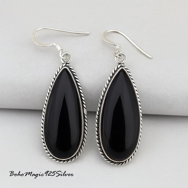 May include: A pair of teardrop-shaped earrings with polished black stones. The stones are framed by a silver-colored, twisted rope design. The earrings have a simple hook closure. The text "BohoMagic925Silver" is visible at the bottom.