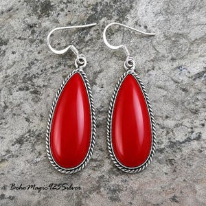 May include: A pair of teardrop-shaped earrings with a vibrant red stone center. The earrings have a silver-colored, twisted rope-like border and a hook closure. The background is a textured, gray surface. The text "Boho Magic 925 Silver" is visible.
