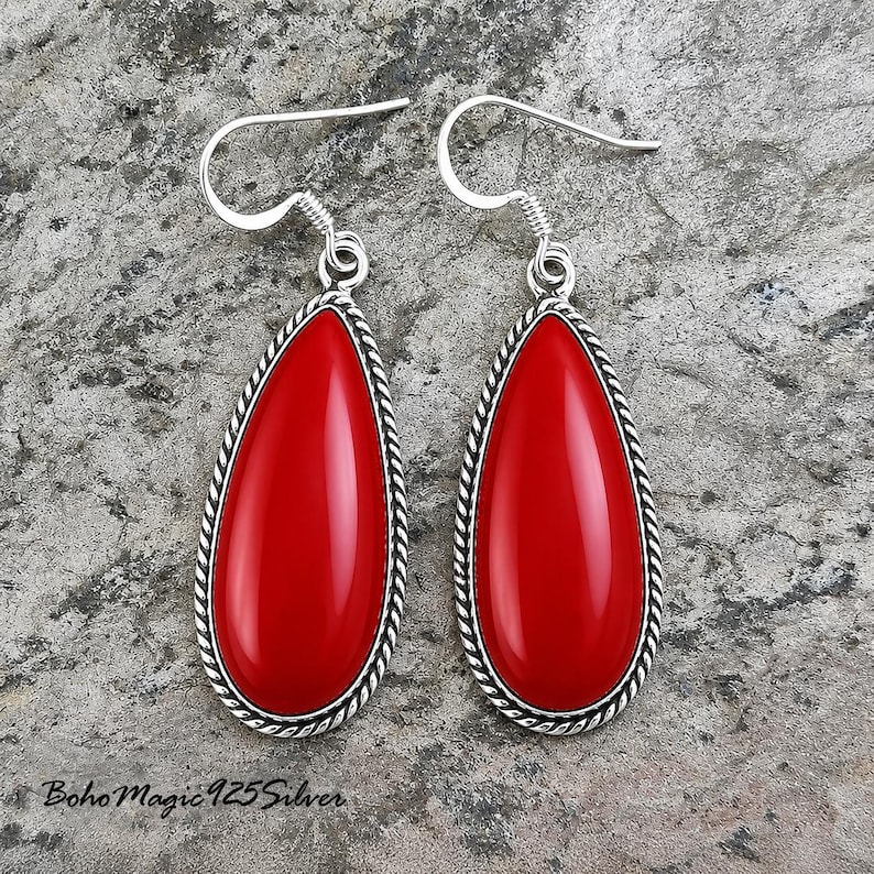 May include: A pair of teardrop-shaped earrings with a vibrant red stone center. The earrings have a silver-colored, twisted rope-like border and a hook closure. The background is a textured, gray surface. The text "Boho Magic 925 Silver" is visible.
