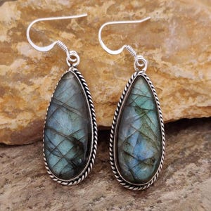 May include: A pair of teardrop-shaped earrings featuring iridescent blue-green labradorite stones. Each stone is framed with a silver-colored, twisted rope design and has a silver hook for pierced ears. The earrings are displayed on a textured, tan surface.