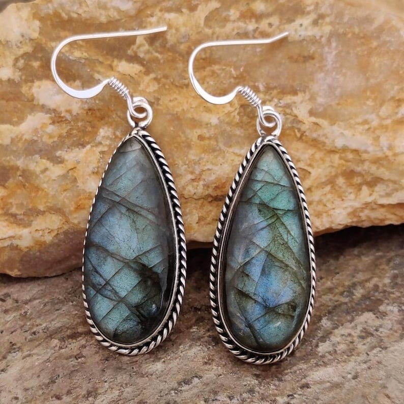 May include: A pair of teardrop-shaped earrings featuring iridescent blue-green labradorite stones. Each stone is framed with a silver-colored, twisted rope design and has a silver hook for pierced ears. The earrings are displayed on a textured, tan surface.