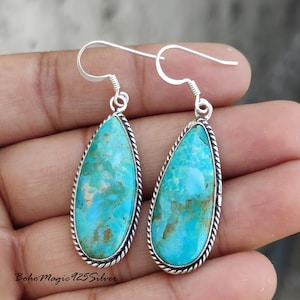 May include: A pair of turquoise teardrop earrings with a silver rope-like border. The earrings have a bright blue and green color with a silver hook. The earrings are held in a person's hand.