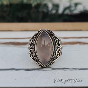 May include: A silver ring featuring a light purple oval gemstone. The ring has an intricate, filigree pattern around the stone and on the band. The ring is displayed on a white and grey surface. The text "BohoMagic925Silver" is visible.