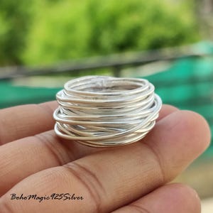 Könnte beinhalten: Ein Silberring mit einem einzigartigen Design. Der Ring besteht aus mehreren Strängen Silberdraht, die gewickelt und aufgerollt sind, um einen strukturierten, geschichteten Effekt zu erzeugen. Der Ring wird in einer Hand gehalten, mit einem verschwommenen grünen Hintergrund.