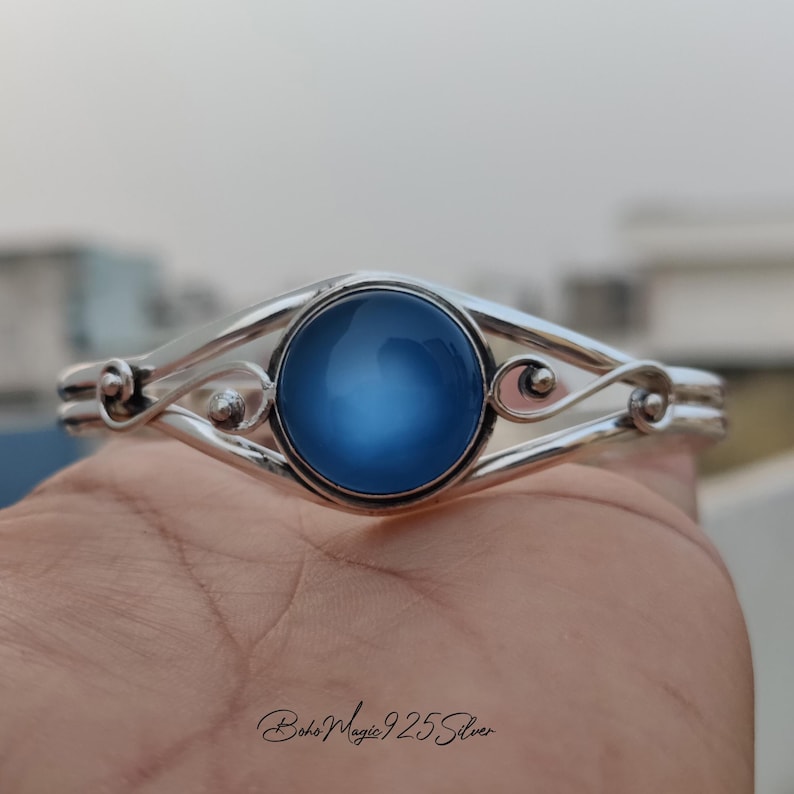 May include: A silver bangle bracelet with a blue cabochon gemstone set in the center. The bracelet has a delicate, swirling design.